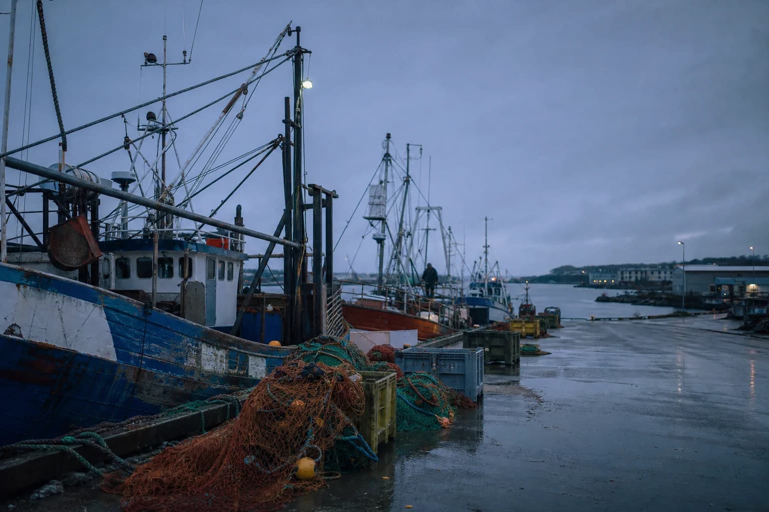 A harbor at first light, small trawlers moored in a row, nets stacked on the quay under a gray sky.